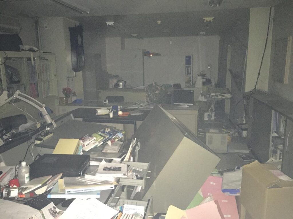 地震で激しく散らかった事務室の内部。大きなキャビネットが倒れ、書類やファイルが床一面に崩れ落ちている