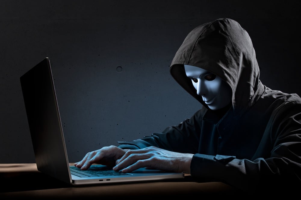 黒いフードを被り白い仮面をつけた人物が、暗い部屋でノートパソコンを操作している写真。サイバー犯罪や匿名性のあるインターネット上の脅威を象徴するイメージです。